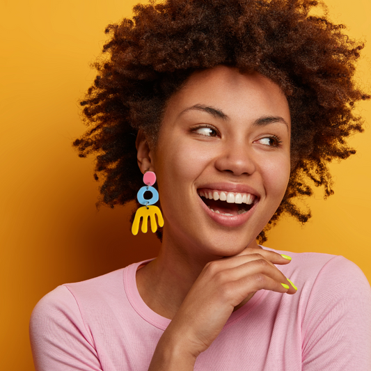lady wearing colourful bright Phat Poly earrings and smiling to the right. The earrings are pink, baby blue and mustard and she is wearing a complimentary pink top.